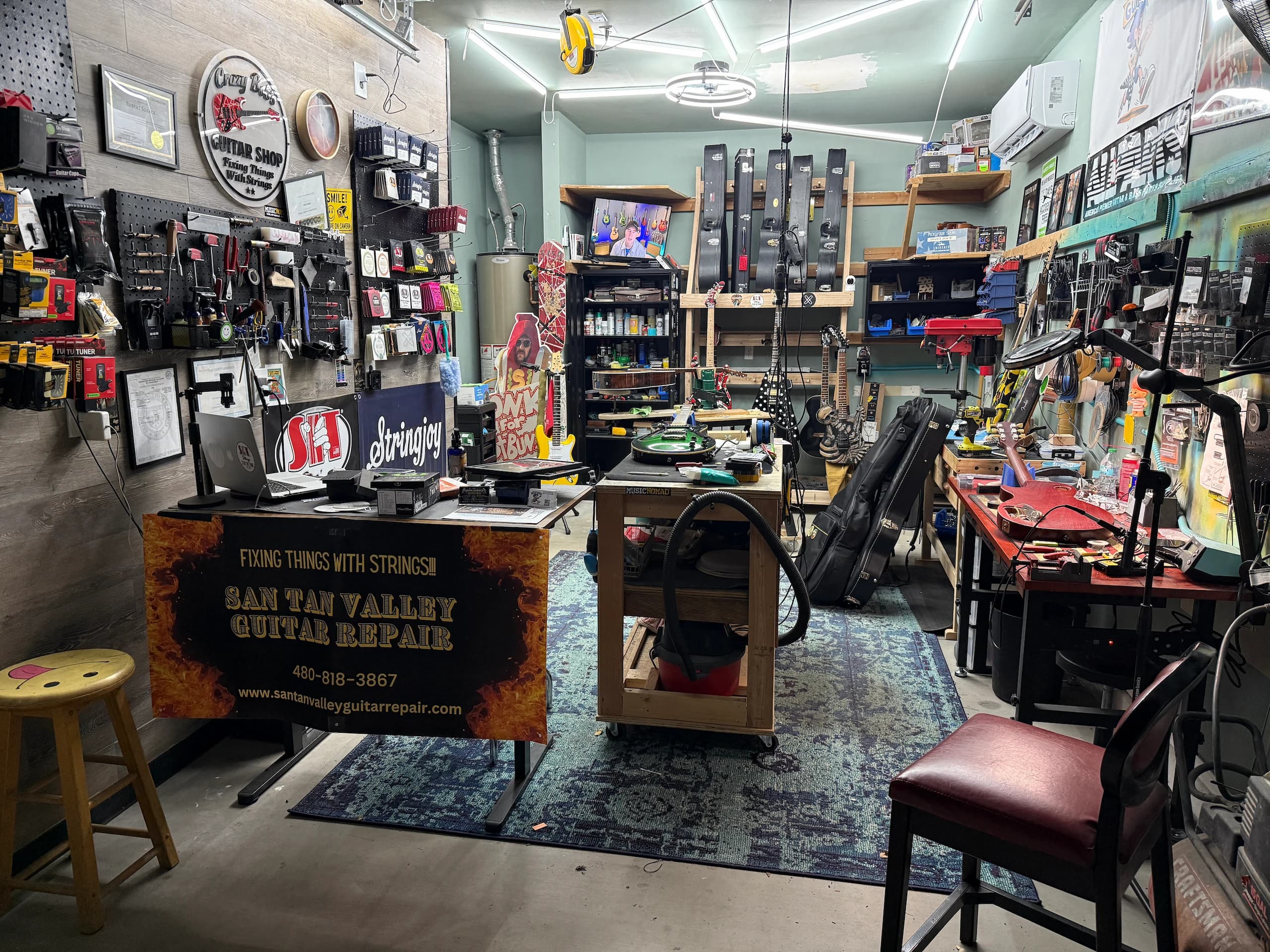 Guitar repair workshop with tool-lined pegboard walls, workbenches holding guitars in progress, instrument cases, and posters.