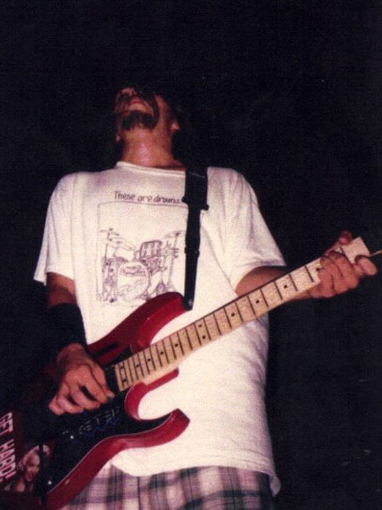 Bob playing a red electric guitar on stage, head tilted back, wearing a white graphic t-shirt.